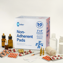 A box of Globe Sterile Non-Adherent Pads (50-pack, 3” x 8”, highly absorbent, non-stick wound dressings) sits on a white surface among medicine bottles, blister packs, ampoules, a rolled bandage, and a white bottle.