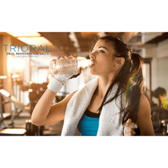 A woman in workout gear drinks water at the gym, a white towel around her neck and exercise equipment behind her. Sunlight streams in. In the corner, the text reads: “TRIORAL Oral Rehydration Salts (15 Packets/Box) by TRIORAL.”.