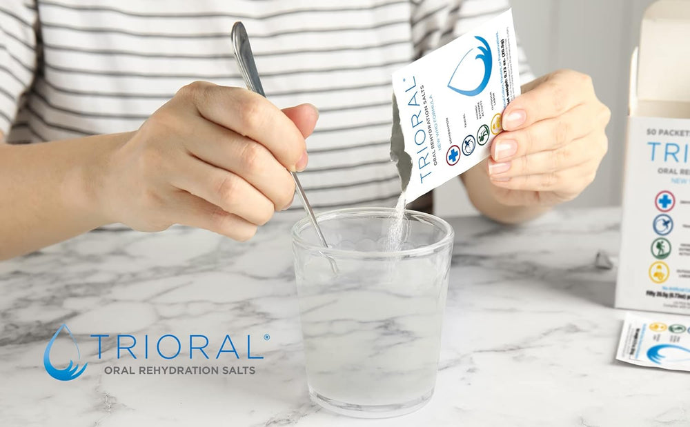 A person stirs a glass of water while adding a TRIORAL Rehydration Electrolyte Powder packet. The TRIORAL box and 100-pack WHO Hydration Supplement Salts Formula are on the marble surface.