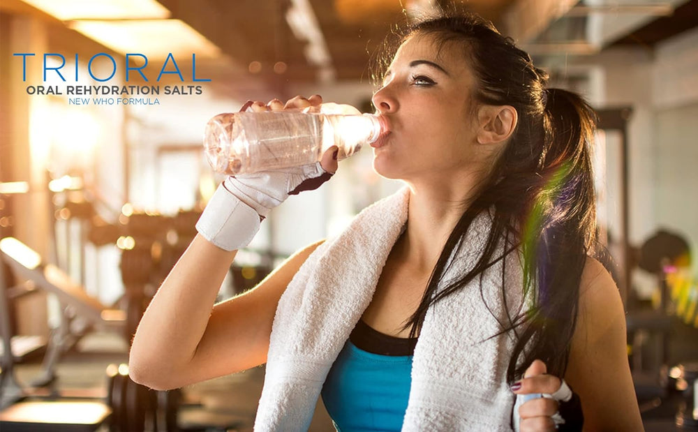 A woman in workout attire drinks from a bottle at the gym, gym equipment visible. The text "TRIORAL Rehydration Electrolyte Powder - WHO Hydration Supplement Salts Formula" appears at top left, highlighting its hydration benefits.