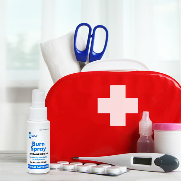 A red first aid kit sits on a table by a window with scissors, gauze, thermometer, medicine bottle, tablets, and Globe Burn Spray—Lidocaine 2% Topical Anesthetic Pain Relief & Numbing 2 oz Spray Bottle from Globe.