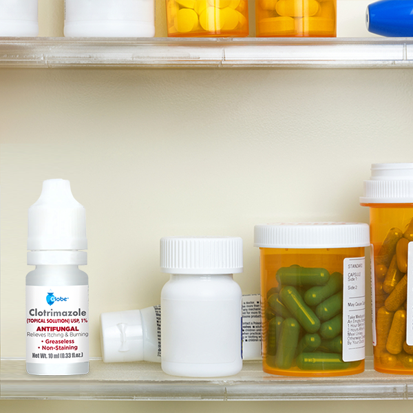 A medicine cabinet shelf holds prescription pill bottles, capsules, and a 10ml bottle of Globe Clotrimazole Solution for Athlete’s Foot, Jock Itch, and Ringworm in the foreground.