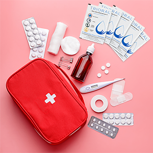 A red first aid kit with white cross is surrounded by pills, bandages, safety pins, cotton, a brown medicine bottle, and a carton of 600 TRIORAL Oral Rehydration Salts sachets—ideal for distributor orders—on a pink background.