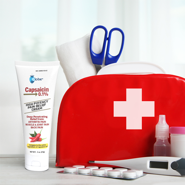 A red first aid kit with medical supplies—including scissors, a thermometer, pills, and Globe Capsaicin 0.1% High Potency Pain Relief Cream (2 oz) for arthritis, muscle, joint, and back pain—sits on a table.