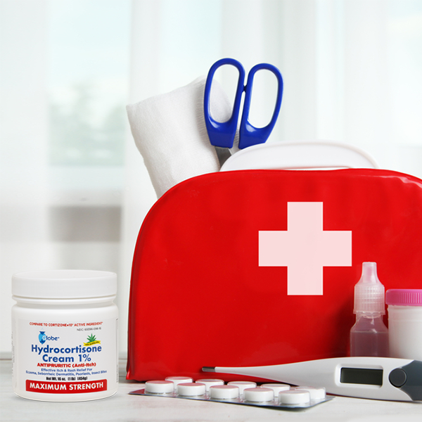 A red first aid kit with a white cross holds scissors, gauze, and medical supplies. In front are Globe Hydrocortisone Maximum Strength Cream 1% w/ Aloe (16 oz), a thermometer, blister pack pills, and a medicine bottle.
