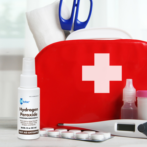 A red first aid kit with a white cross holds scissors, gauze, and supplies. In front is a Globe Hydrogen Peroxide 3% First Aid Antiseptic Spray Bottle (2 fl. oz), plus a digital thermometer, blister-pack pills, and medicine bottles.