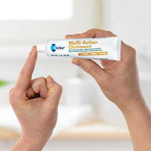 A person dispenses Globe Multi-Action Ointment 1 oz onto their fingertip. This first aid ointment treats minor cuts, scrapes, burns and relieves pain and itching against a blurred indoor background.