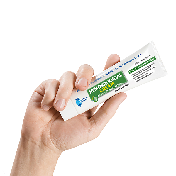 A hand holds a tube of Globe Hemorrhoid Symptom Treatment Cream (1.8 oz), for pain relief with aloe, featuring white, green, and blue labeling by Globe, against a plain white background.