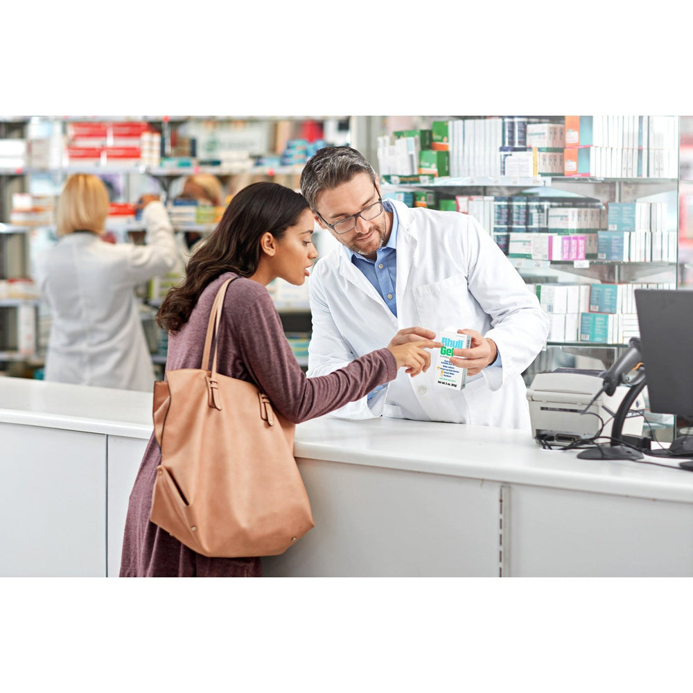 A pharmacist hands a prescription bag to a woman at the counter while explaining medication instructions, including information about Rhuli Gel Anti-Itch Gel for relief from bug bites and skin irritation; shelves of medicine are visible in the background.