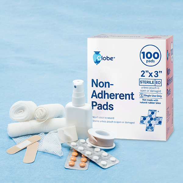 A box of Globe Sterile Non-Adherent Pads 2" x 3" sits on a blue surface, surrounded by gauze rolls, adhesive bandages, pill blister packs, tape, mesh, and a white spray bottle. These pads are highly absorbent, non-stick, and individually wrapped.