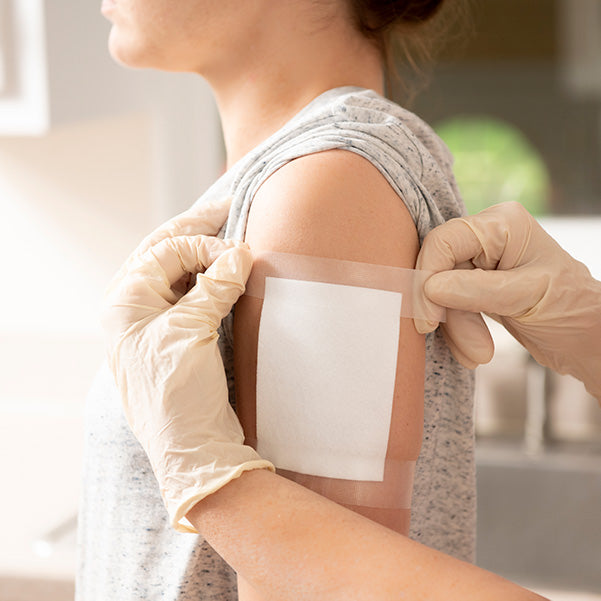 Someone in a grey shirt receives a Globe Sterile Non-Adherent Pad (3” x 4”, 100-Pack) on their upper arm from a gloved person, showing the importance of Globe’s highly absorbent, painless removal first aid essentials.