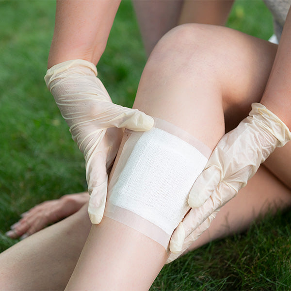 A person wearing latex gloves applies Globe 4” x 4” Sterile Gauze Pads as a wound dressing to another’s knee while sitting outdoors on grass. The highly absorbent, 12-ply cotton pads are individually packed and ideal for home first aid kits.