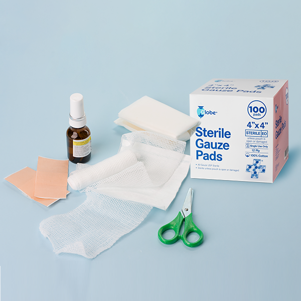 First aid supplies on a light blue background, featuring Globe 4"x4" Sterile Gauze Pads (100-pack), a spray bottle, adhesive bandages, wound dressing rolls, folded gauze pads, and green-handled scissors.